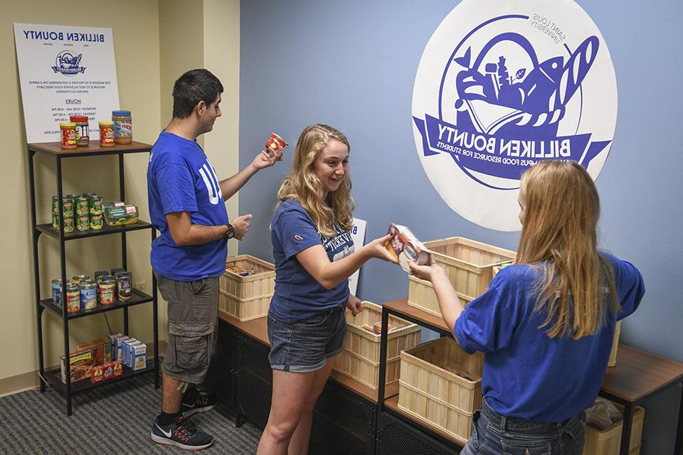 Three students examine packages of food amongst baskets and shelves of donated goods.