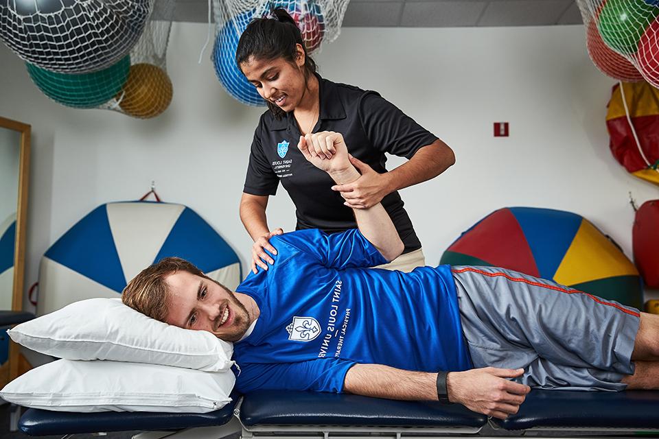 A physical therapy student assists a patient lying on a table with an arm exercise, with physical therapy equipment in the background.