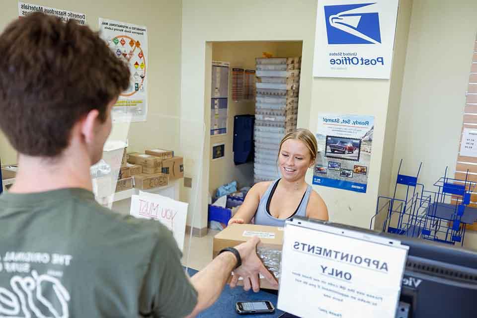 A mailroom worker surrounded by packages hands a student (seen from the back) his mail
