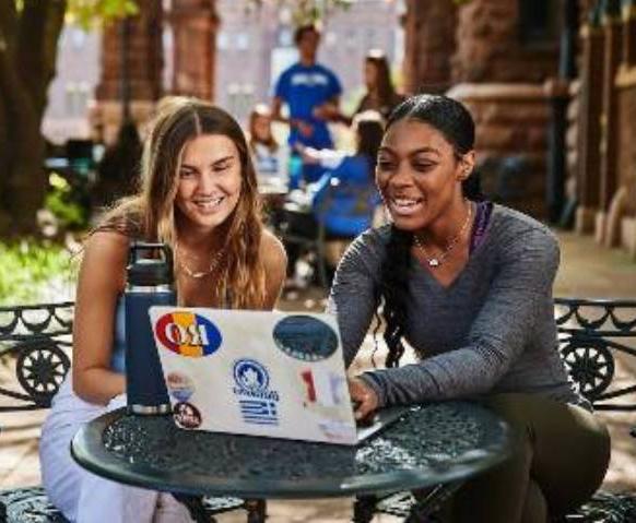 Two smiling students sitting outdoors at a small cafe table studying with laptops in front