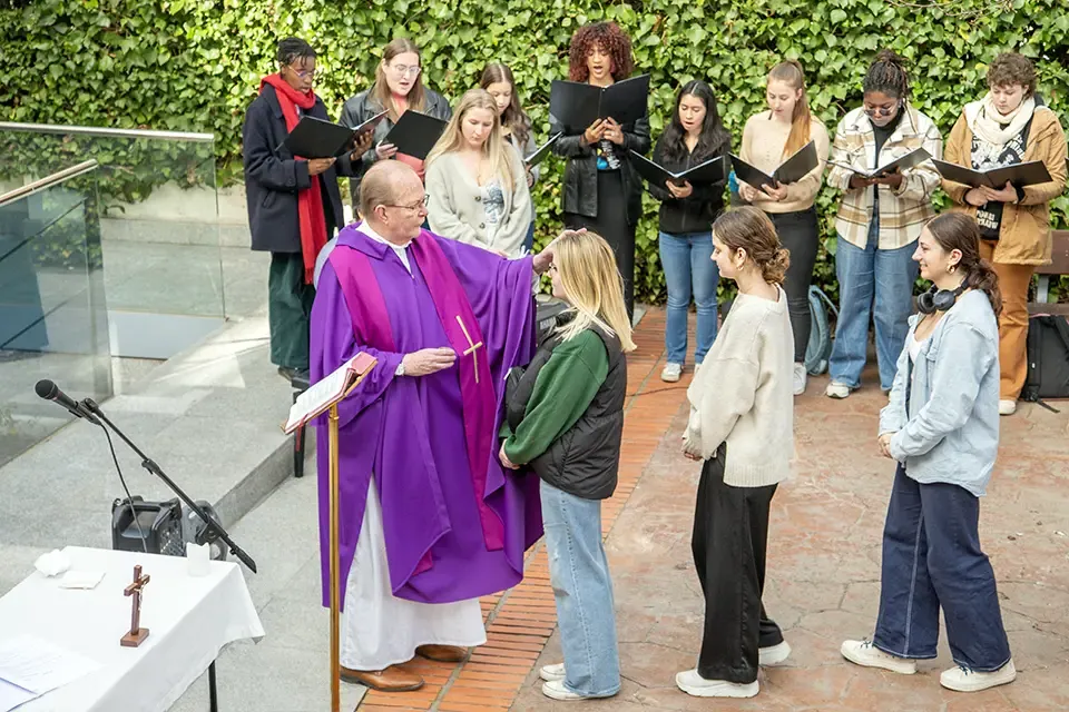 Students line up in an outdoor area while a priest marks a student's forehead. A choir of students stands nearby holding black choir books and singing.
