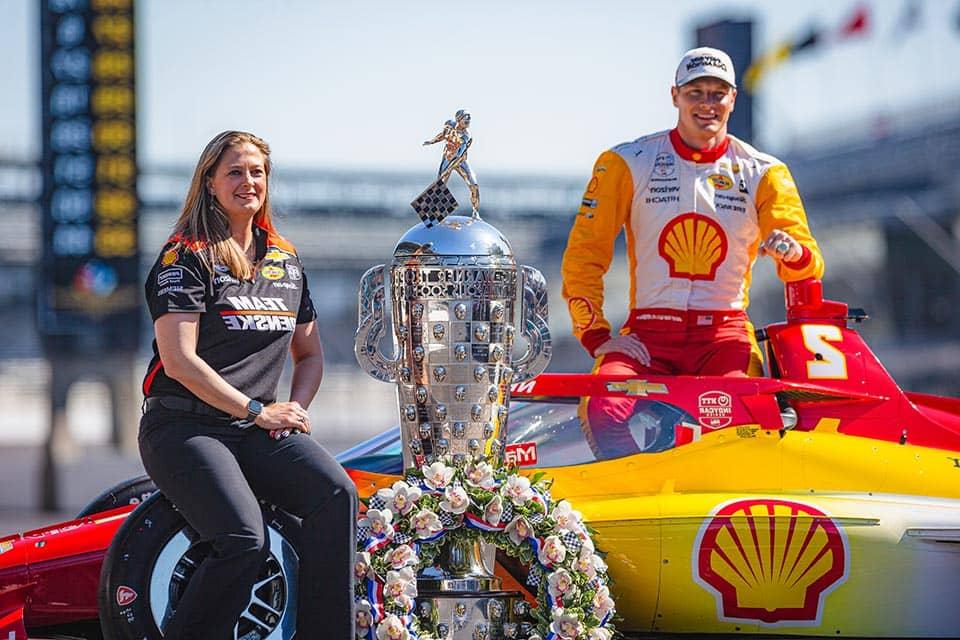 Sullivan sits in front of a racecar near a trophy won at the Indy 500 across from a driver.